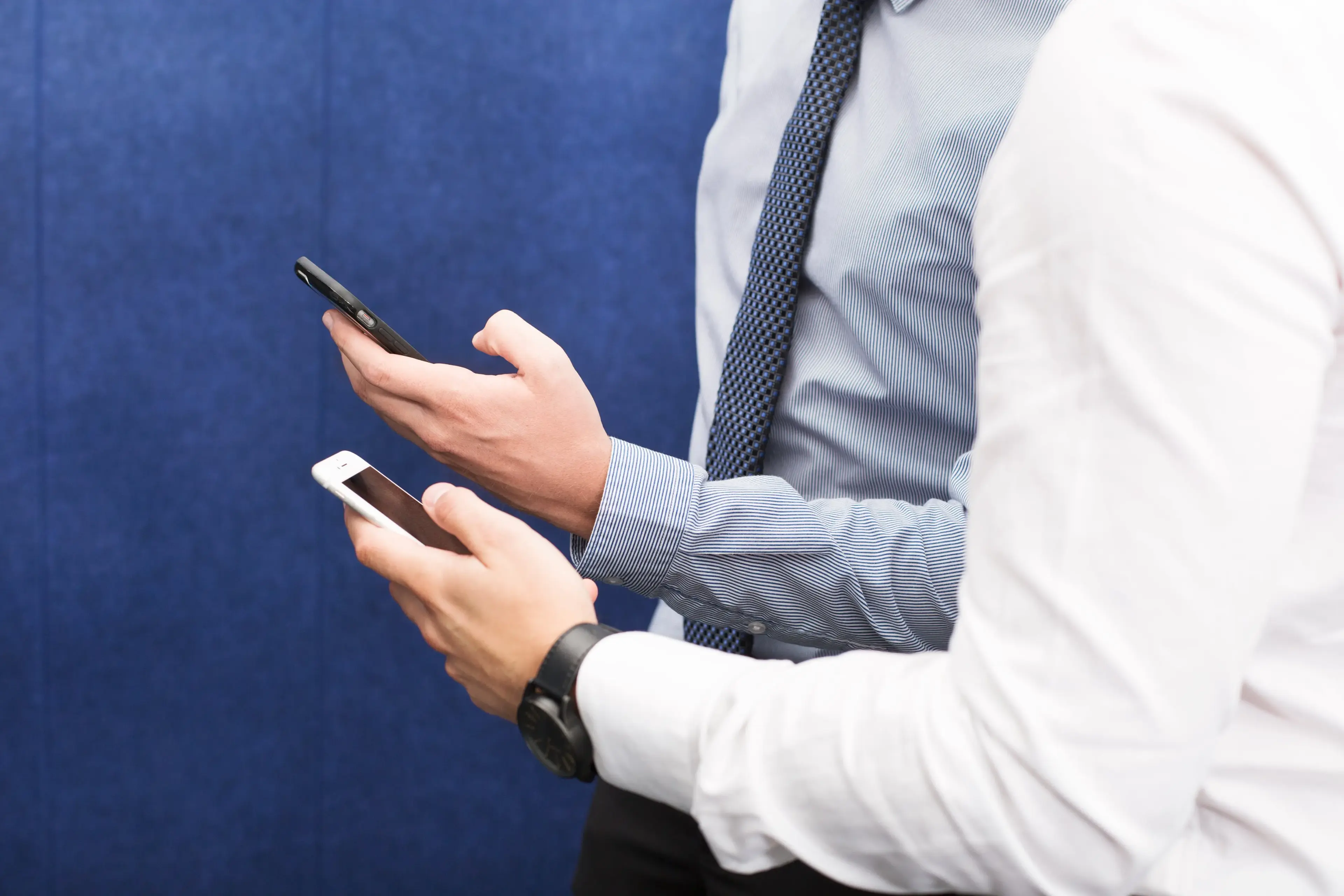 Two people wearing office shirts both hold mobile phones with their hands and type on them