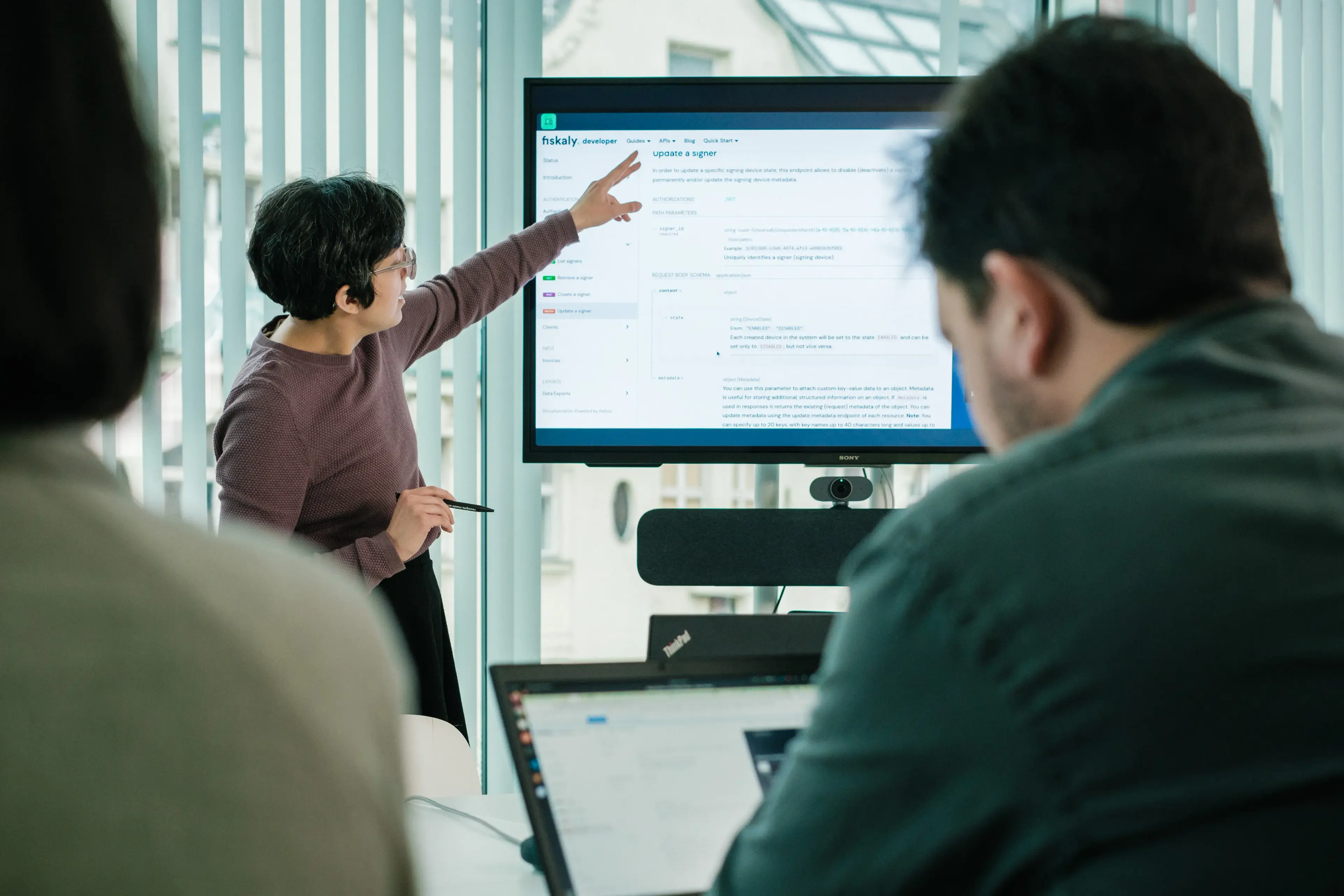 In the background, there is a woman, showing a fiskaly documentation page for KassenSichV on the big monitor.